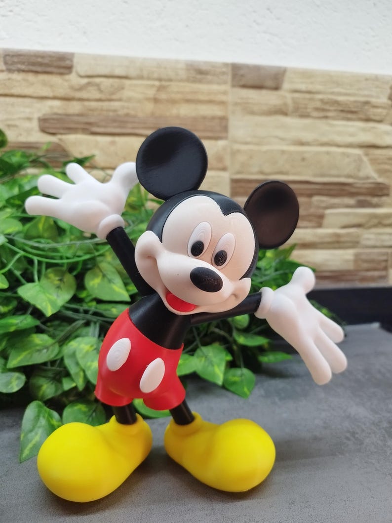 May include: A figurine of Mickey Mouse, a classic cartoon character, is depicted with outstretched white gloved hands. Mickey wears red shorts with white buttons, yellow shoes, and a black body and ears. The figurine is set against a backdrop of green leaves and a textured wall.