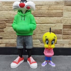 Könnte beinhalten: Zwei Zeichentrickfiguren, Sylvester the Cat und Tweety Bird, sind in Freizeitkleidung dargestellt. Sylvester trägt einen grünen Kapuzenpullover, graue Shorts und rote Turnschuhe. Tweety trägt einen rosa Kapuzenpullover, blaue Shorts und lila Turnschuhe. Die Figuren stehen auf einer grauen Oberfläche.