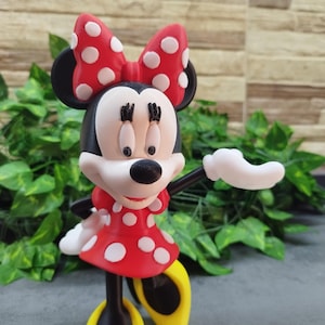 May include: A Minnie Mouse figurine, featuring a red dress with white polka dots and a matching bow. The figure has black ears, a white face, and yellow shoes. The figurine is posed with one arm extended.