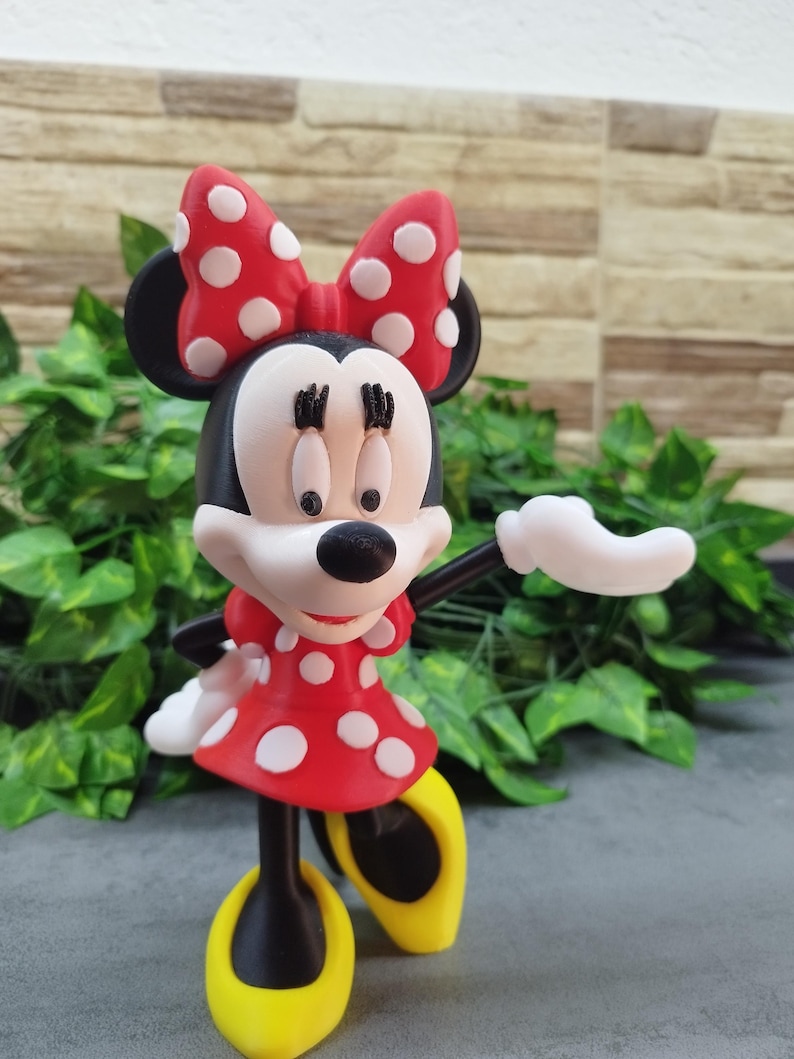 May include: A Minnie Mouse figurine, featuring a red dress with white polka dots and a matching bow. The figure has black ears, a white face, and yellow shoes. The figurine is posed with one arm extended.