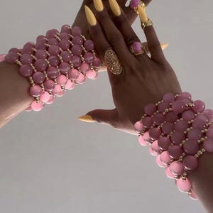 May include: Two hands adorned with yellow nail polish and gold rings display pink beaded bracelets. The bracelets feature large, round beads in varying shades of pink, connected by gold-colored spacers. The rings include a filigree design and pink gemstones.