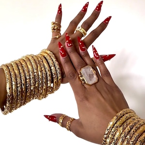 May include: Gold-toned jewelry set featuring multiple bangles with etched designs, rings, and a large ring with a rectangular stone. The nails are painted red with gold accents. The jewelry is displayed against a white background.