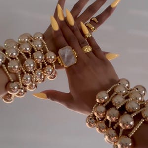 May include: A display of gold jewelry, including multiple rings and bracelets. The bracelets feature large, round beads. The nails are painted a bright yellow. A large, rectangular ring with a light-colored stone is also visible.