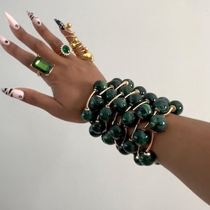 May include: A hand adorned with gold rings and a bracelet featuring dark green beads. The rings include a large emerald-cut green stone and a spiral design. The bracelet has multiple strands of dark green beads connected by gold-colored metal.