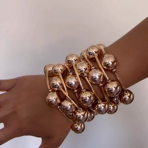Puede incluir: Pulseras doradas con una serie de grandes cuentas esféricas conectadas por barras curvas. Las pulseras están apiladas en una muñeca, mostrando su superficie pulida y reflectante. La joyería tiene un diseño audaz y moderno.
