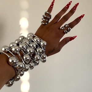 May include: A silver-toned bracelet with multiple spherical beads and matching rings. The rings include a spiral design and a cluster of small spheres. The nails are painted red with gold accents. The jewelry is displayed against a white background.
