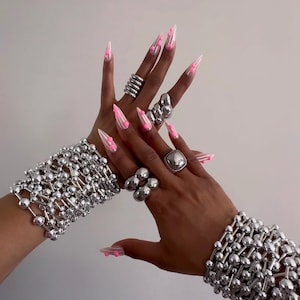 Può includere: Mani adornate con gioielli d'argento e unghie lunghe e appuntite con disegni rosa e bianchi. I gioielli includono anelli e bracciali fatti di sfere collegate. Lo sfondo è di colore chiaro.
