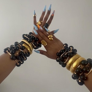 May include: The image displays gold rings and bracelets. The bracelets combine black beads with gold elements. The nails are painted light blue with nail art. The jewellery is set against a plain white backdrop.