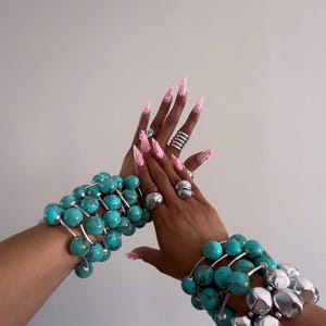 May include: Two turquoise beaded bracelets with silver accents are shown. The image also displays multiple silver rings and long pink and white manicured nails. The bracelets are made of large, round turquoise beads strung together with silver wire.