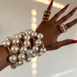 May include: A collection of gold jewelry and a pearl bracelet. The bracelet features large, round, white pearls strung together with gold-colored metal. The hand also displays multiple gold rings, including a thick, stacked ring and a ring with pearl accents.