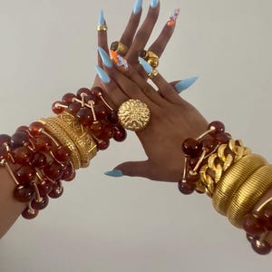 May include: A collection of gold rings and bracelets, including amber-coloured bead bracelets and a large gold ring with a sunburst design. The nails are painted light blue with some accent nails featuring orange and white designs.