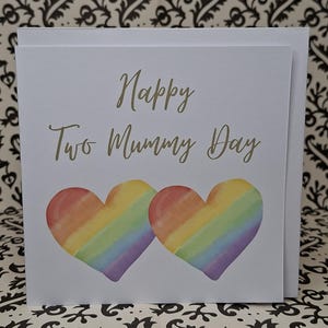 May include: A white greeting card with the text "Happy Two Mummy Day" in gold script. Two watercolor rainbow hearts are below the text. The card is on a white background.