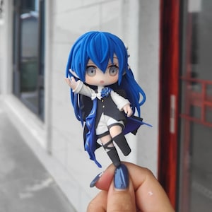 Op de afbeelding: Een miniatuur anime-stijl figuur met helderblauw haar en ogen. Het figuur draagt een zwart-witte outfit met blauwe accenten, waaronder een cape en een bloemdetail. Het figuur wordt tegen een vage buitenachtergrond gehouden.