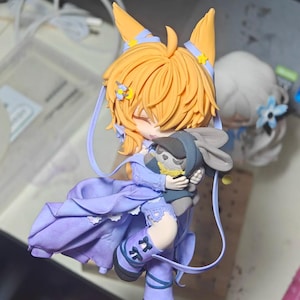 Op de afbeelding: Handgemaakte figuur van een personage met oranje haar en vossenoortjes, gekleed in een paarse jurk en laarzen. De figuur houdt een kleine grijze konijn vast. Het beeldje heeft een gedetailleerd ontwerp met bloemaccenten.