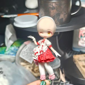 Op de afbeelding: Een kleine, handgemaakte pop met een knipoog. De pop draagt een rood-witte jurk met een gerimpelde rok, witte handschoenen en roze schoenen. De pop heeft een kale kop en gele ogen. Op de achtergrond een koffiemok en knutselspullen.