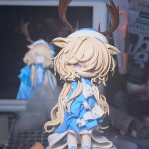 Op de afbeelding: Gedetailleerd beeldje van een personage met lang, lichtblond haar, paarse ogen en bruine geweien. Het personage draagt een blauwe jurk met witte accenten en houdt een klein wit knuffeldier vast. Het beeldje staat op een ronde, grijze basis.