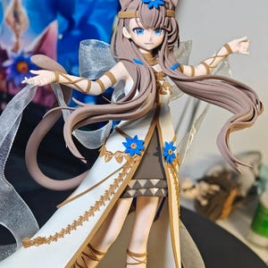 Op de afbeelding: Gedetailleerd beeldje van een vrouwelijk anime-personage met kattenoren, blauwe ogen en lang bruin haar. Ze draagt een witte en gouden jurk met blauwe bloemaccenten. Het beeldje heeft een vloeiend, elegant ontwerp met een lint en een staart.