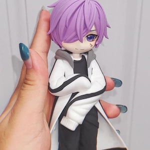 Op de afbeelding: Een handgemaakt beeldje van een anime-stijl karakter met paars haar en blauwe ogen. Het figuur draagt een witte jas met zwarte bies over een zwart shirt en broek. Het karakter heeft een klein teken onder één oog.