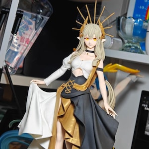 Op de afbeelding: Gedetailleerd beeldje van een vrouwelijk personage met lang blond haar, gele ogen en puntige oren. Ze draagt een witte top, een zwarte en gouden jurk en een gouden hoofdtooi. Het beeldje poseert met een vloeiende witte stof.