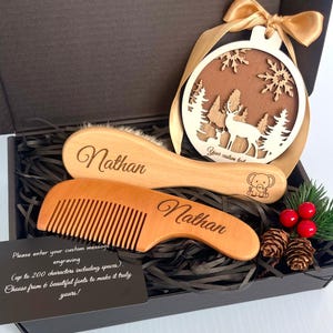May include: A wooden baby brush and comb set engraved with "Nathan", a Christmas ornament with a deer and tree design, and a gift box. The ornament has a gold ribbon. A card in the box says "Please enter your custom message engraving".