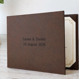 May include: A dark brown leather certificate holder featuring the engraved text Emma & Daniel 24 August 2026 on the front cover. The folder is partially open, revealing a cream-coloured document with a decorative gold border held by corner tabs.