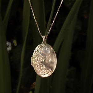 Può includere: Un ciondolo a medaglione ovale in argento sospeso a una sottile catena a maglie quadrate. Il medaglione presenta un motivo floreale in rilievo che copre il lato sinistro della superficie, mentre la parte restante è liscia e lucida.