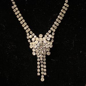 Puede incluir: Un collar plateado con un grupo central de brillantes redondos y transparentes. El collar tiene un diseño en cascada con múltiples hilos de brillantes más pequeños, creando un aspecto brillante y elegante. El fondo es negro.