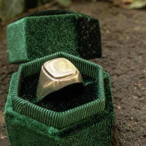 May include: A silver signet ring with a smooth, square-shaped face featuring rounded corners. The ring is displayed inside an open, hexagonal-shaped dark green velvet jewellery box with a matching lid resting behind it.
