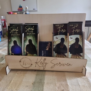 May include: A wooden display stand holding several books with dark covers. The books have titles like "Spark Your Imagination" and "Light Your Fire." The stand is light-colored with the name "Kelly Scriven" engraved on the front.