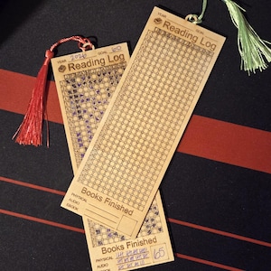 May include: Two wooden reading log bookmarks with tassels. One bookmark shows a goal of 60 books for the year 2026, with some squares filled in. The other bookmark shows 65 books finished. The bookmarks are on a black and red surface.