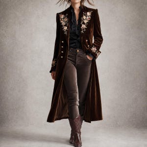May include: A long brown velvet coat featuring double-breasted gold buttons and floral embroidery on the lapels and cuffs. The coat is worn over a black button-down shirt and dark brown trousers, paired with brown leather western-style boots.