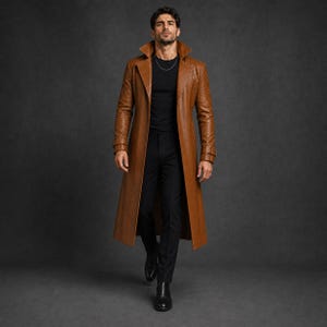 May include: A long tan leather trench coat with a wide collar and belted cuffs, worn over a black crew neck t-shirt and black trousers. The coat features a structured silhouette, long sleeves, and a smooth finish, paired with black leather boots.