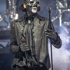 May include: A performer in a black leather jacket, black shirt, and black pants, wearing a silver skull mask and black gloves. The performer is holding a microphone and a microphone stand. The background is blurred with stage lights.