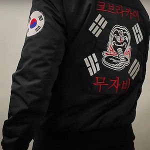 Könnte beinhalten: Schwarze Bomberjacke mit einem Aufnäher mit einer Kobra und Text in Koreanisch und Japanisch. Die Jacke hat auch einen Aufnäher mit der südkoreanischen Flagge auf dem linken Ärmel. Ein lässiges Oberbekleidungsstück.