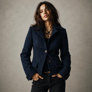 May include: A dark navy blue blazer featuring a notched lapel, two front buttons, and two flap pockets at the waist. The blazer has long sleeves with buttoned cuffs and is worn over a black top paired with black trousers, creating a structured look.