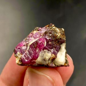 Puede incluir: Primer plano de una gema de rubí en bruto. El rubí es de un rojo intenso y vibrante con estructuras cristalinas visibles. Está engastado en una matriz de minerales blancos y marrones, creando un contraste llamativo. La gema se sostiene en una mano.