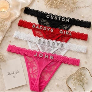 Peut inclure: Un ensemble de quatre strings en dentelle noire, rouge, blanche et rose, chacun doté d'une ceinture festonnée avec des lettres en strass. Le string noir porte l'inscription CUSTOM, le rouge DADDYS GIRL, le blanc DADDYS et le rose JOHN.