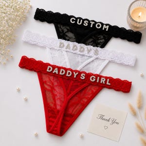 Op de afbeelding: Drie kanten strings in zwart, wit en rood. Elk paar heeft een geschulpte tailleband versierd met strass-letters. De zwarte string heeft de tekst CUSTOM, de witte DADDYS en de rode string heeft de tekst DADDYS GIRL.