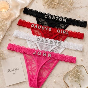 May include: Four pairs of lace thong underwear in black, red, white, and hot pink. Each pair features a scalloped lace waistband adorned with silver-toned rhinestone letters spelling out CUSTOM, DADDYS GIRL, DADDYS, and JOHN respectively.