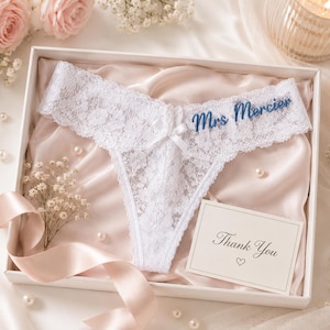 May include: White lace thong featuring a small center bow and the embroidered blue text Mrs Mercier. The garment is presented in a gift box lined with pale pink satin, accompanied by a white card with the printed text Thank You and a heart icon.