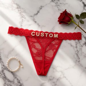 Peut inclure: String en dentelle rouge avec une ceinture festonnée ornée du mot CUSTOM en lettres dorées incrustées de strass. Le vêtement est accompagné d'un bracelet de perles blanches et d'une rose rouge foncée unique.