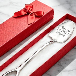 May include: A silver-toned metal ice cream shovel with a spade-shaped handle and a flat, rounded scoop head. The scoop is engraved with the text "Sonny's Ice Cream Shovel." The shovel is presented inside a long, rectangular red gift box with a red bow.