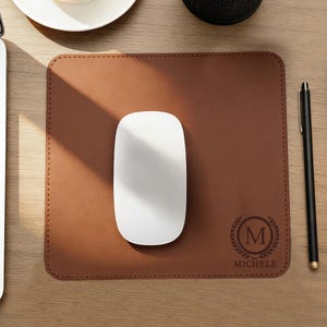 Peut inclure: Un tapis de souris en cuir marron avec une souris blanche, un stylo noir et un bloc-notes sur un bureau en bois. Le tapis de souris a une bordure cousue et un monogramme avec la lettre "M" et le nom "MICHELE". Une tasse de café et un ordinateur portable sont également visibles.