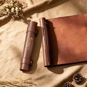 May include: A brown leather portfolio and a matching cylindrical case, both with gold-colored accents. The cylindrical case is embossed with the name "Sonny". The portfolio has a zipper closure. The items are on a tan surface.