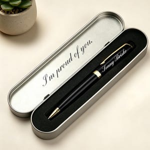 May include: A black pen with gold detailing rests in an open silver case. The pen is inscribed with the name "Sonny Briske." The case is lined with black fabric and features the phrase "I'm proud of you" printed on the inside.