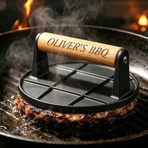 Op de afbeelding: Een zwarte gietijzeren burgerpers met een houten handvat waarop de tekst OLIVER'S BBQ is gegraveerd. De pers staat op een sissende vleesburger in een zwarte grillpan, met zichtbare stoom die van het kookoppervlak opstijgt.