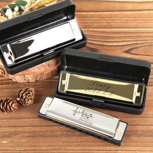 May include: Three harmonicas in black cases on a wooden surface. One harmonica has the name "Sonny" and a musical note design. Another has the name "Fonny" and the date "2022.2.2". The harmonicas are silver and gold.