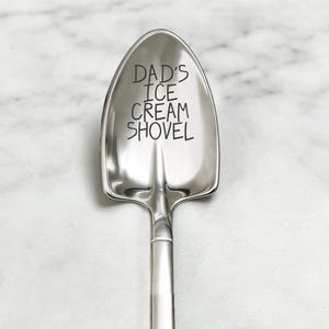 May include: A shiny, silver ice cream shovel with the words "DAD'S ICE CREAM SHOVEL" etched on the scoop. The shovel has a classic spade shape and a long, slender handle. The background is a light-colored, marble-like surface.