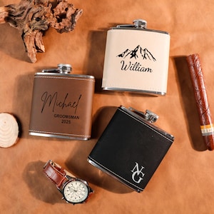 May include: Three personalized flasks in brown, black, and cream colors, with silver accents. The brown flask has the name "Michael" and "Groomsman 2025". The cream flask has a mountain graphic and the name "William". A watch and cigar are also present.