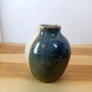 May include: A small, rounded ceramic vase featuring a two-tone design. One side is a smooth, off-white cream colour, while the other side displays a textured, mottled glaze in shades of deep blue, forest green, and earthy brown with a glossy finish.